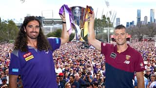 A Fremantle player and a Brisbane Lions player hold the AFL premiership cup above their heads in front of a large crowd in Melbourne.