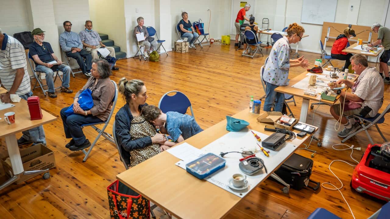People sit on chairs in a large room with tables covered in electronic equipment.
