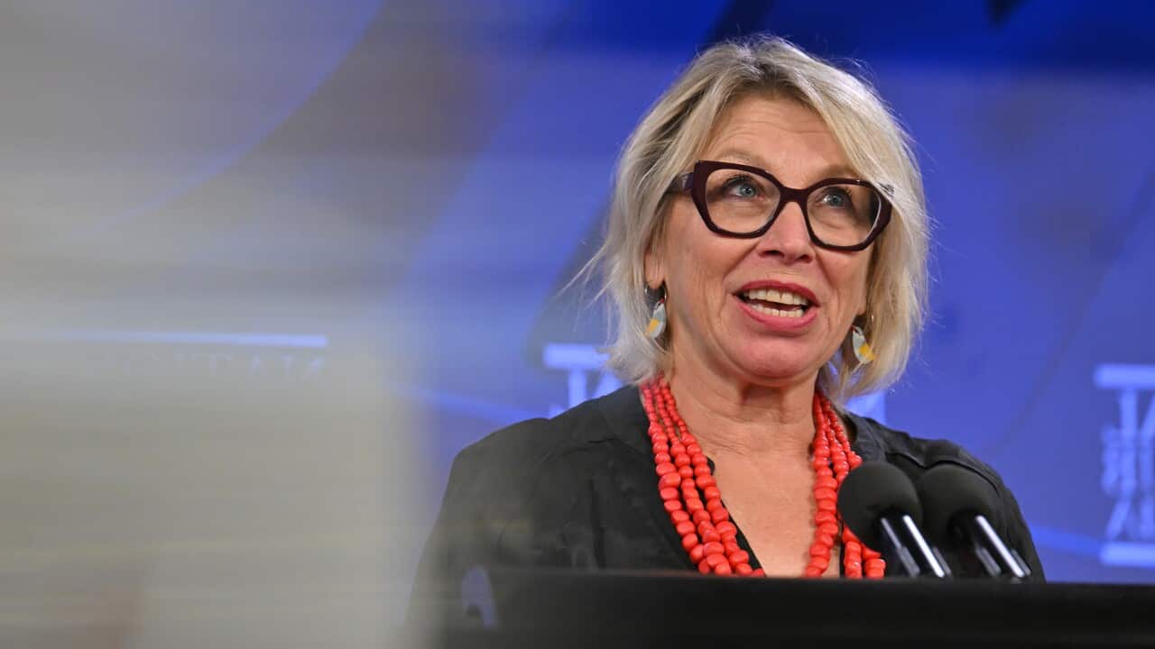 A woman in a black top, wearing glasses and red beads around her neck, is speaking at a podium in front of microphones.