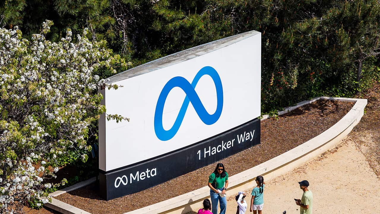 A group of six people stand on a dusty path, close to a large white sign with a blue, looped logo.