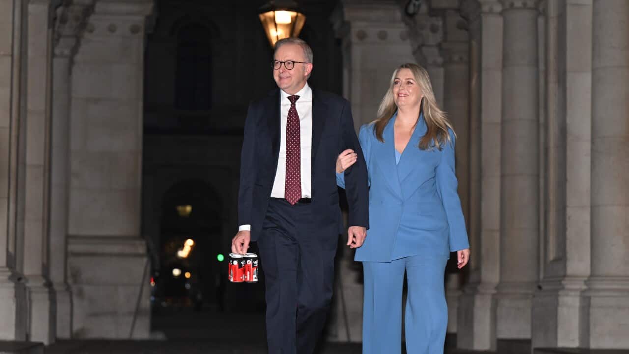 A well-dressed couple walking outside a building. The woman is wearing a bright blue pantsuit, and the man is in a dark suit, holding a six-pack of beer cans.