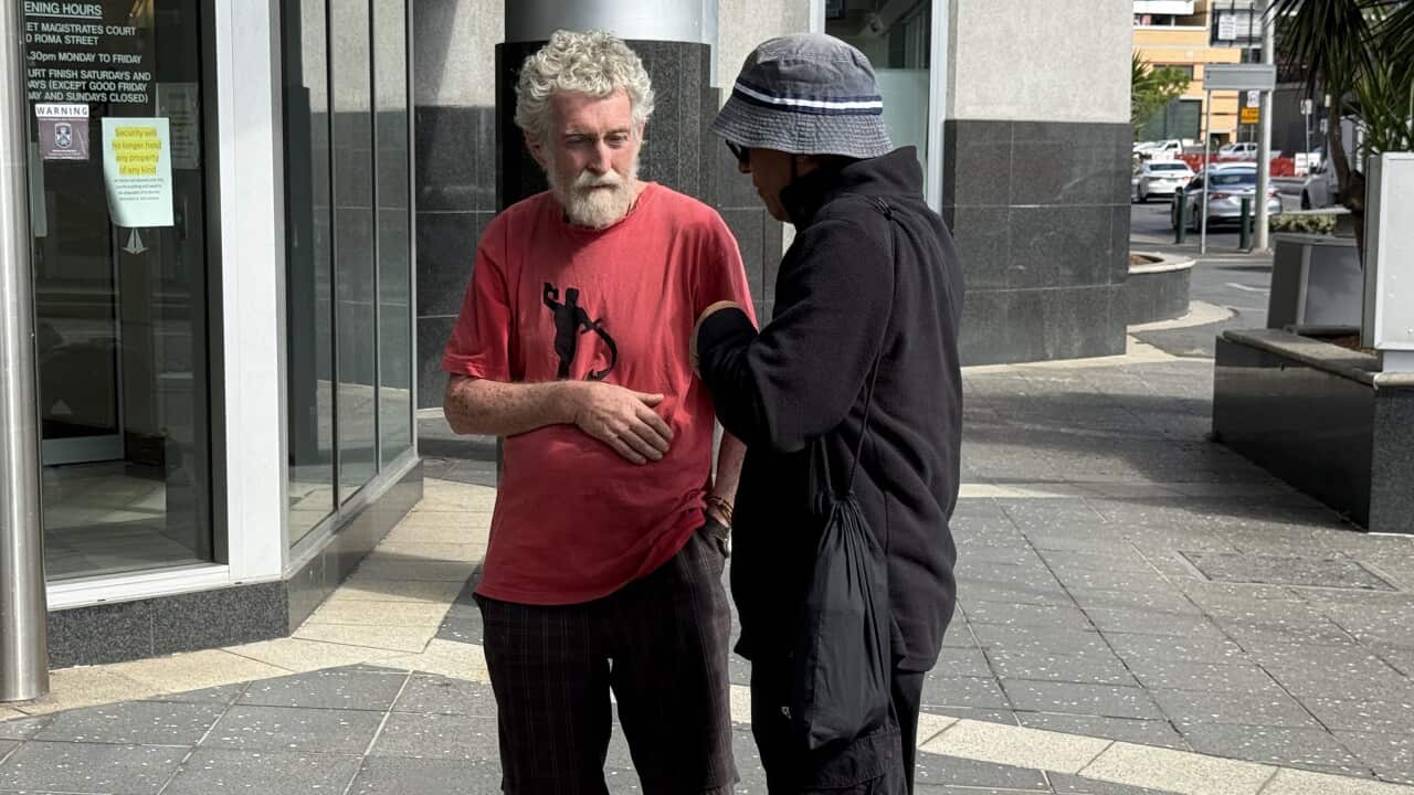 Two men, one in a red t-shirt and barefoot and the other in dark clothing and a bucket hat, stand talking on a city sidewalk outside a building entrance.