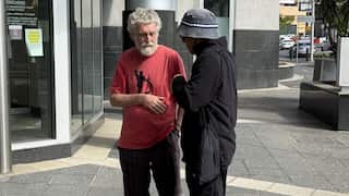 Two men, one in a red t-shirt and barefoot and the other in dark clothing and a bucket hat, stand talking on a city sidewalk outside a building entrance.