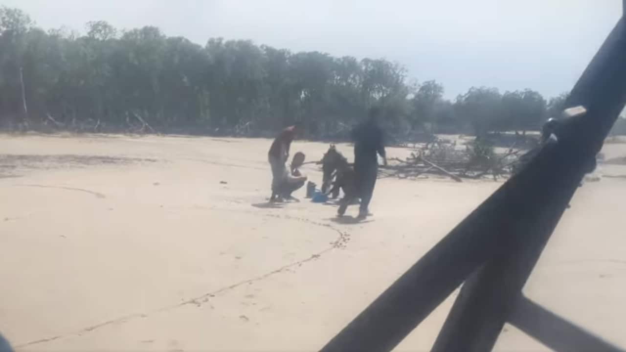A screenshot from a video showing five men standing and squatting on an otherwise deserted beach.