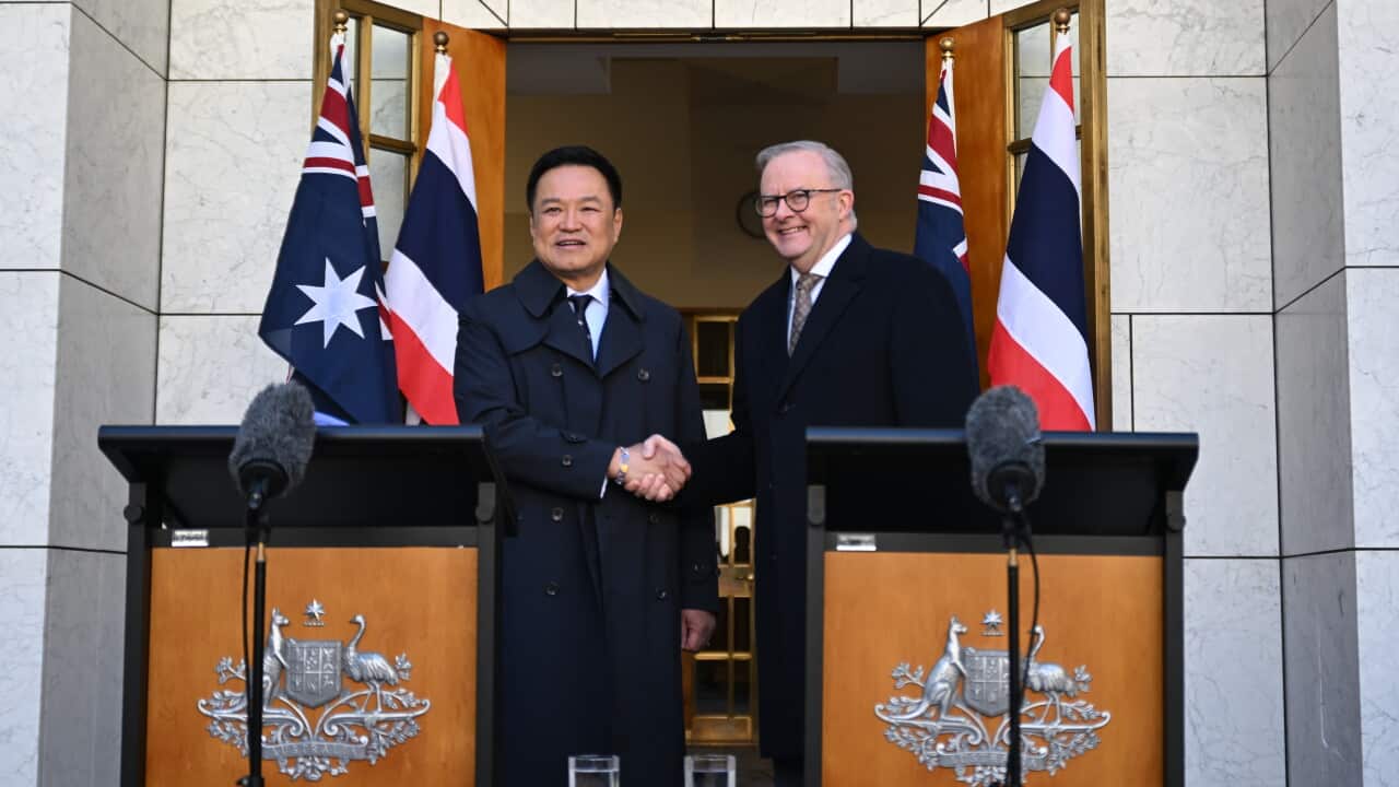 Two men, one middle-aged with dark hair and one older with grey hair, shake hands and smile. They stand behind podiums with microphones in front of a white marble building.