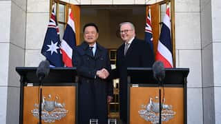 Two men, one middle-aged with dark hair and one older with grey hair, shake hands and smile. They stand behind podiums with microphones in front of a white marble building.