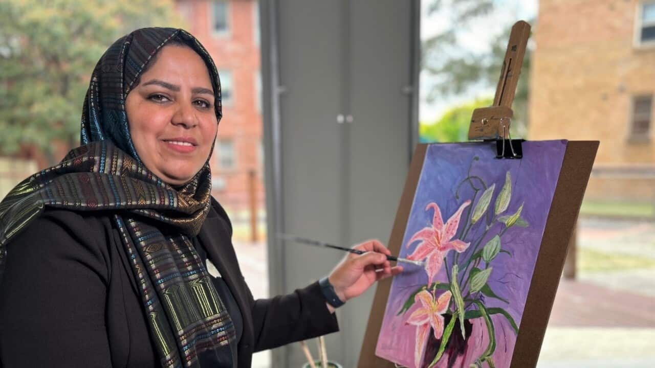 A woman in a brown jacket and headscarf sits at an easel painting a vase of flowers.