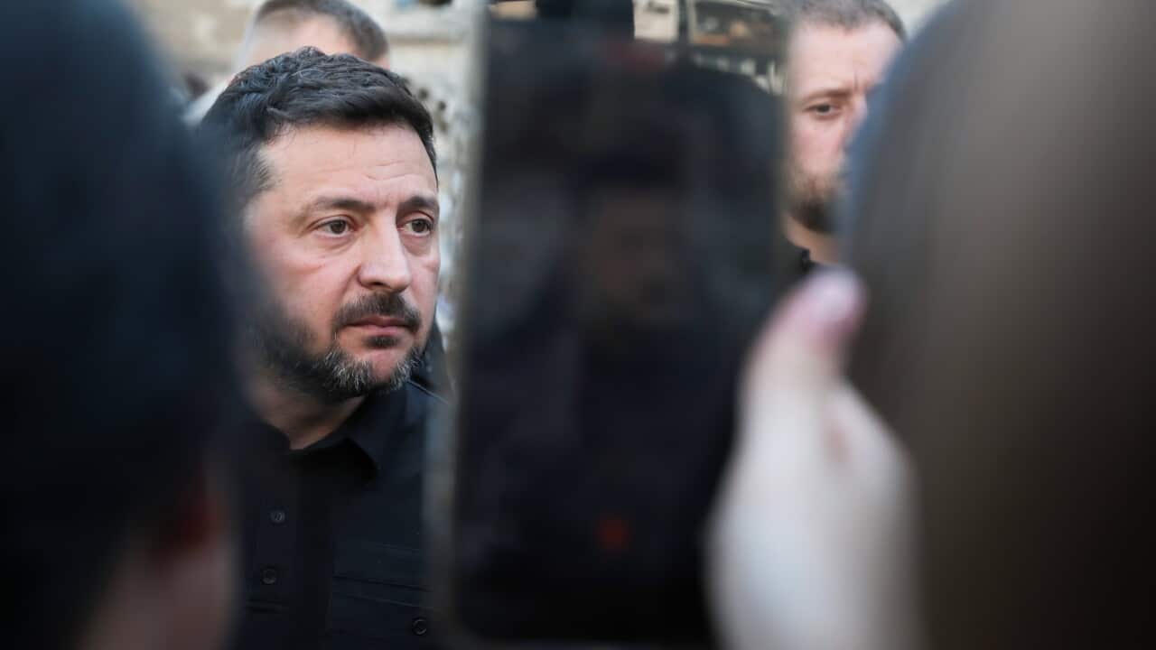 A beareded middle aged man dressed in black is photographed on a smartphone by an onlooker. The man stares ahead, expressionless.