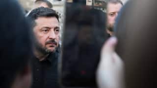 A beareded middle aged man dressed in black is photographed on a smartphone by an onlooker. The man stares ahead, expressionless.