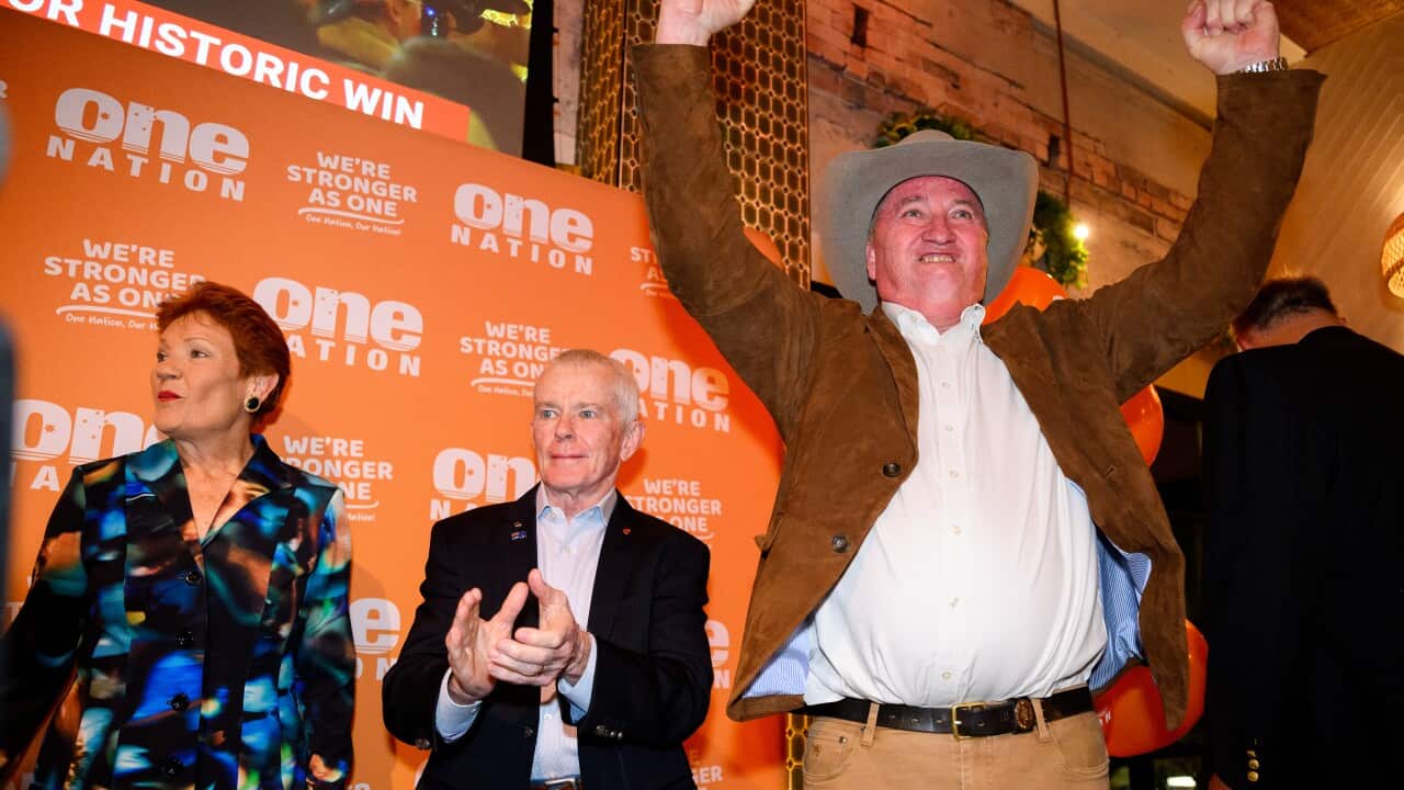 A white man wearing a brown jacket and akubra-style hat raising his arms in celebration. A man and a woman stand beside him.