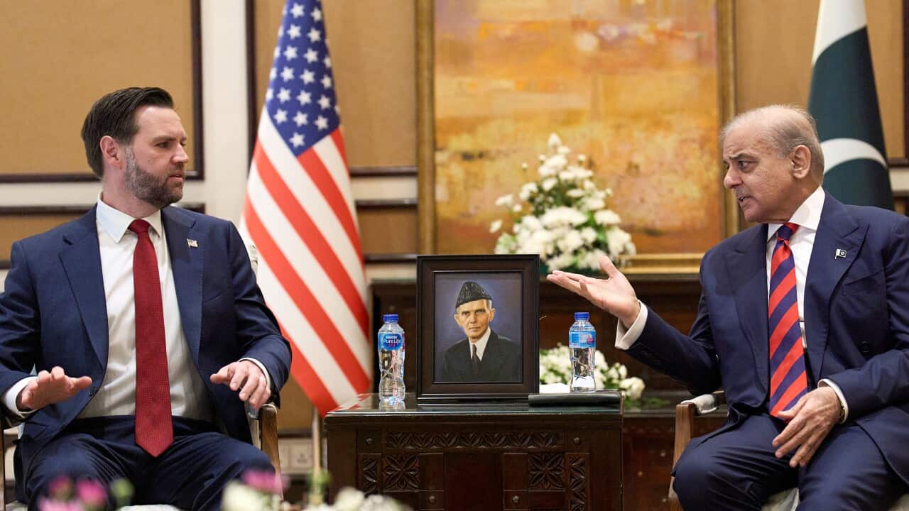 Two men in suits sit in chairs with portrait between them.