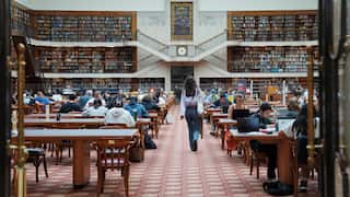 People sit and work at long tables inside a large public library reading room lined with bookshelves.