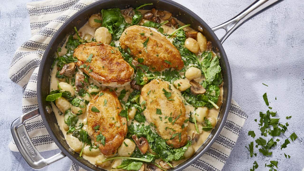 A skillet sits on a striped napkin, on a blue-ish surface. It holds a bake with four golden cooked chicken breasts, gnocchi and greens in a creamy sauce.