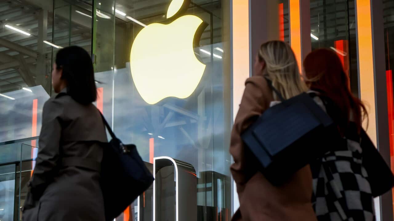 Three women walking past a building. The Apple logo is illuminated on the signage.