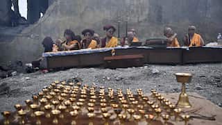 Buddhist monks sitting on the ground near rubble. Some gold objects are in the foreground.