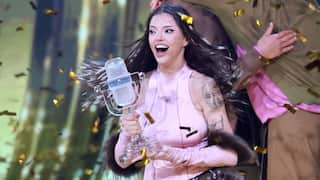 A smiling dark haired woman in a pink top accepts an award as confetti sprays around her.