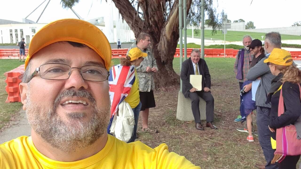 A bespectacled man in a yellow shirt and cap poses in front of a crowd.