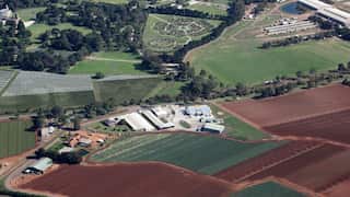 Aerial view of a large agricultural property with crop fields, farm buildings, access roads and surrounding parkland, showing a mix of cultivated land and green open spaces.