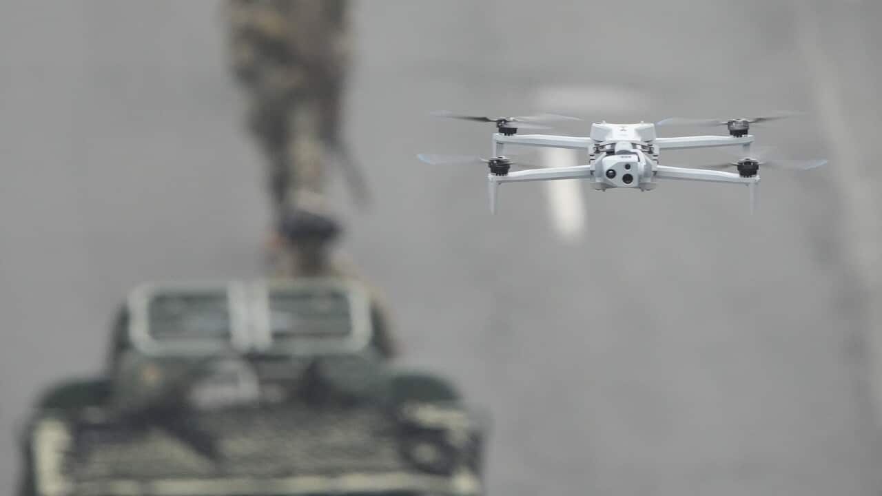 A drone flies over a road with a vehicle and soldiers walking in the background.