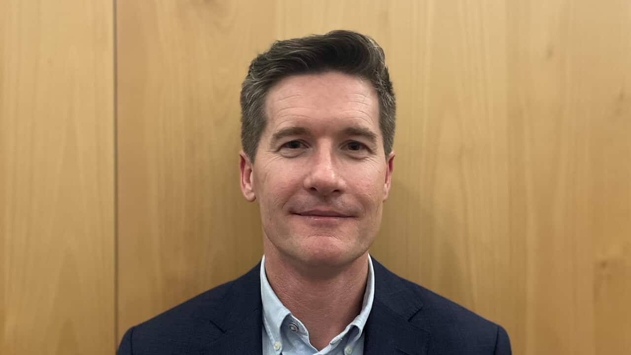 a middle aged man in a suit looks neutrally at camera in front of a plain wooden background
