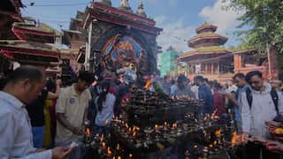 Crowd gathered at a temple square, lighting oil lamps and incense before a large decorated deity statue, with historic multi-tiered buildings surrounding the scene.