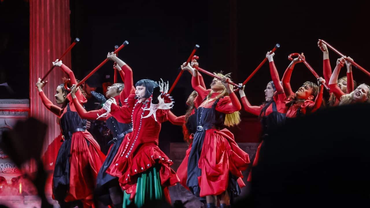 A musician in a red and green gown performs in the centre, along with other dancers in red and black outfits holding red sticks during a concert.