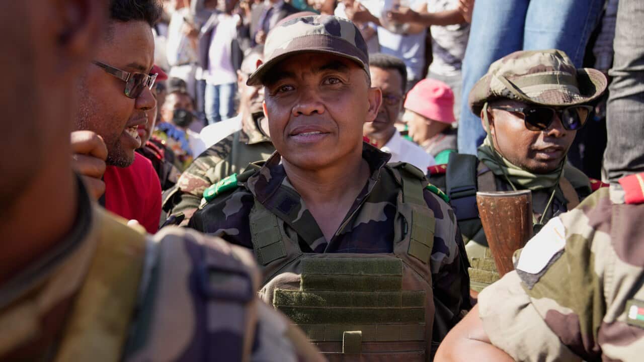 A man in a military uniform is flanked by military personnel, and a crowd is visible behind them.