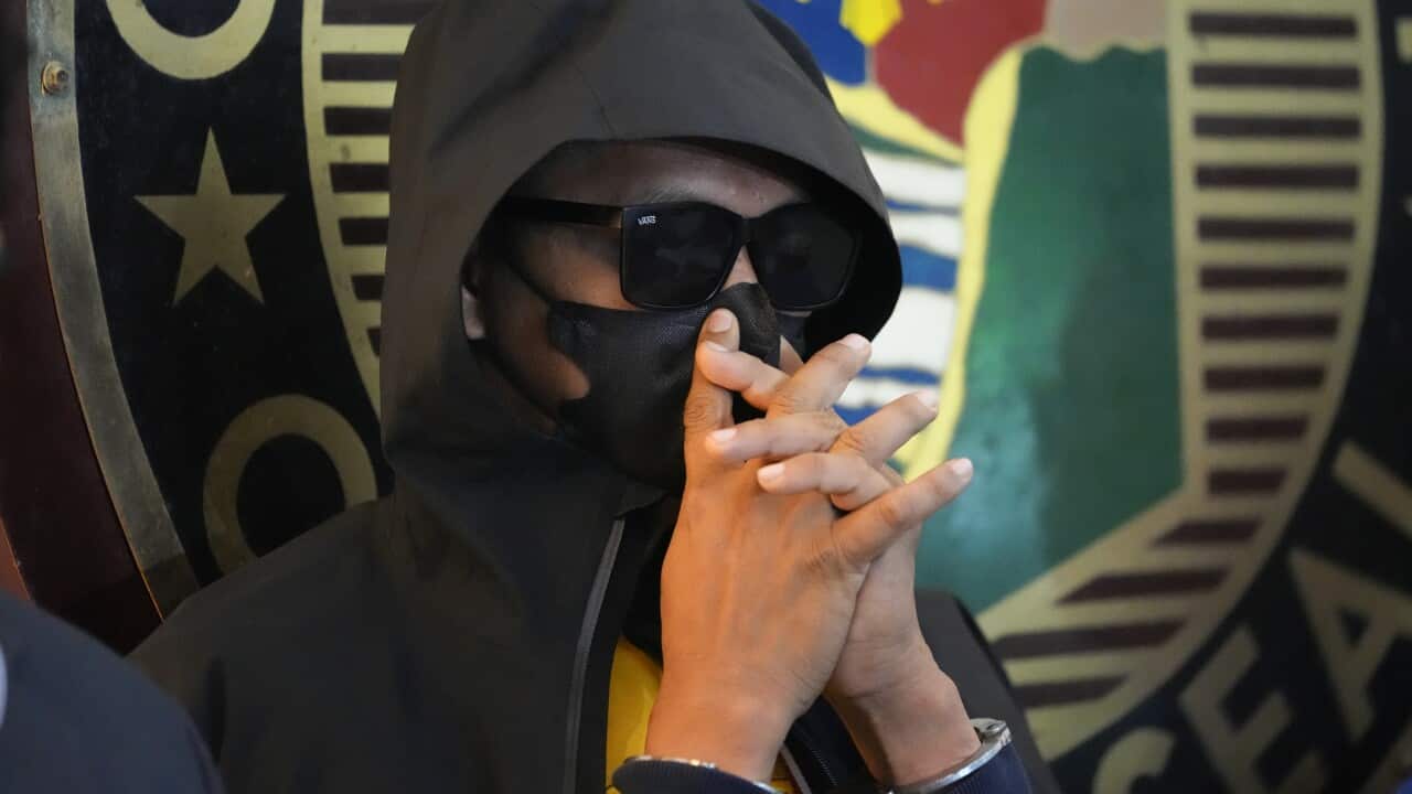 A man with sunglasses, a hood and a face mask, covering his face with his hands.