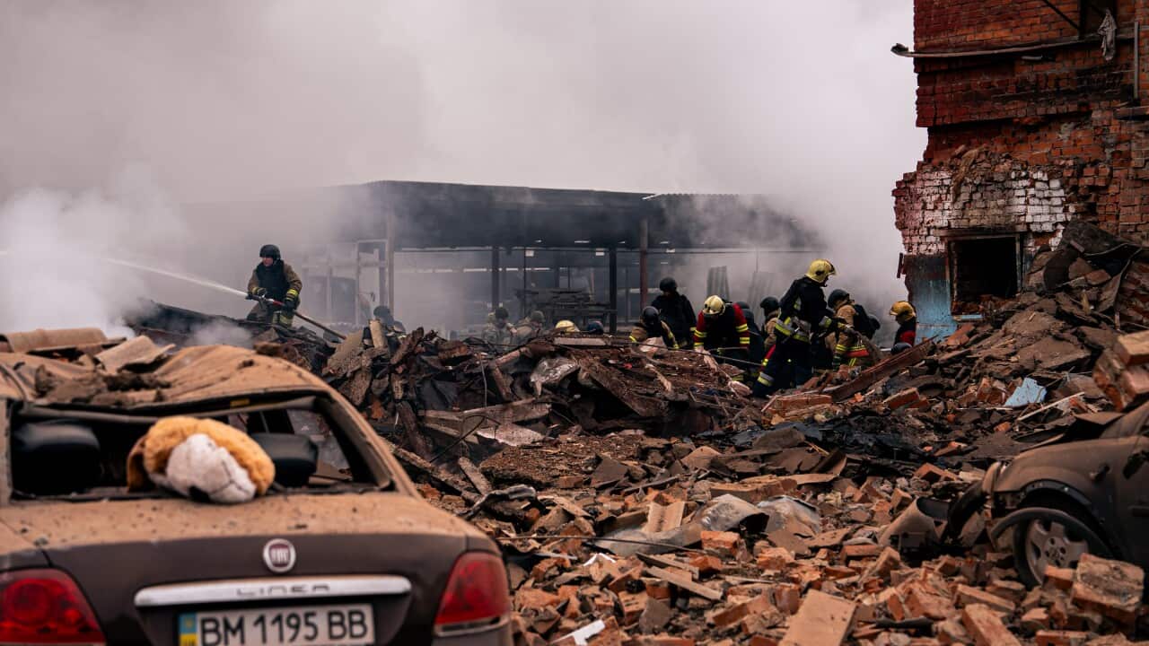 Firefighters extinguish flames and clear debris from the rubble of a collapsed building, with a completely destroyed sedan visible in the foreground.