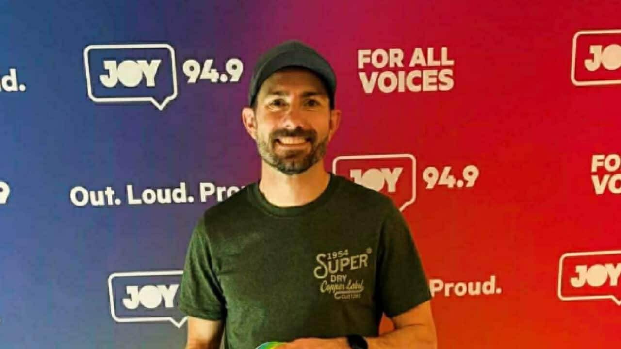 Leigh Ryswyk, the first openly gay AFL player, stands before a rainbow-coloured Joy FM media wall and holds a rainbow-coloured AFL football in his hands. He is a white man in his late thirties, smiling at the camera through a short beard. He wears a green baseball cap and matching green shirt with black shorts.
