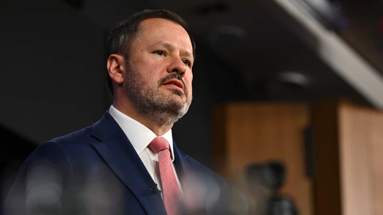 A man in a dark suit and a red tie is speaking into a microphone.
