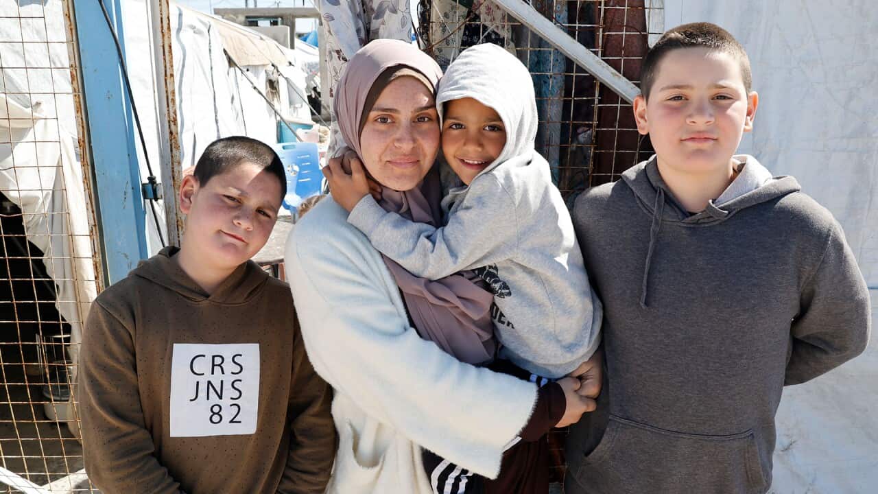 A woman wearing a hijab is holding a young boy in a grey hoodie with his hands wrapped around her neck. Two pre-teen boys are standing on each side of her.