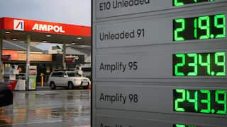 A board of high fuel prices fills the frame, figures as high as 243.9 for Amplify 98. A car sits at a petrol bowser in the background/.