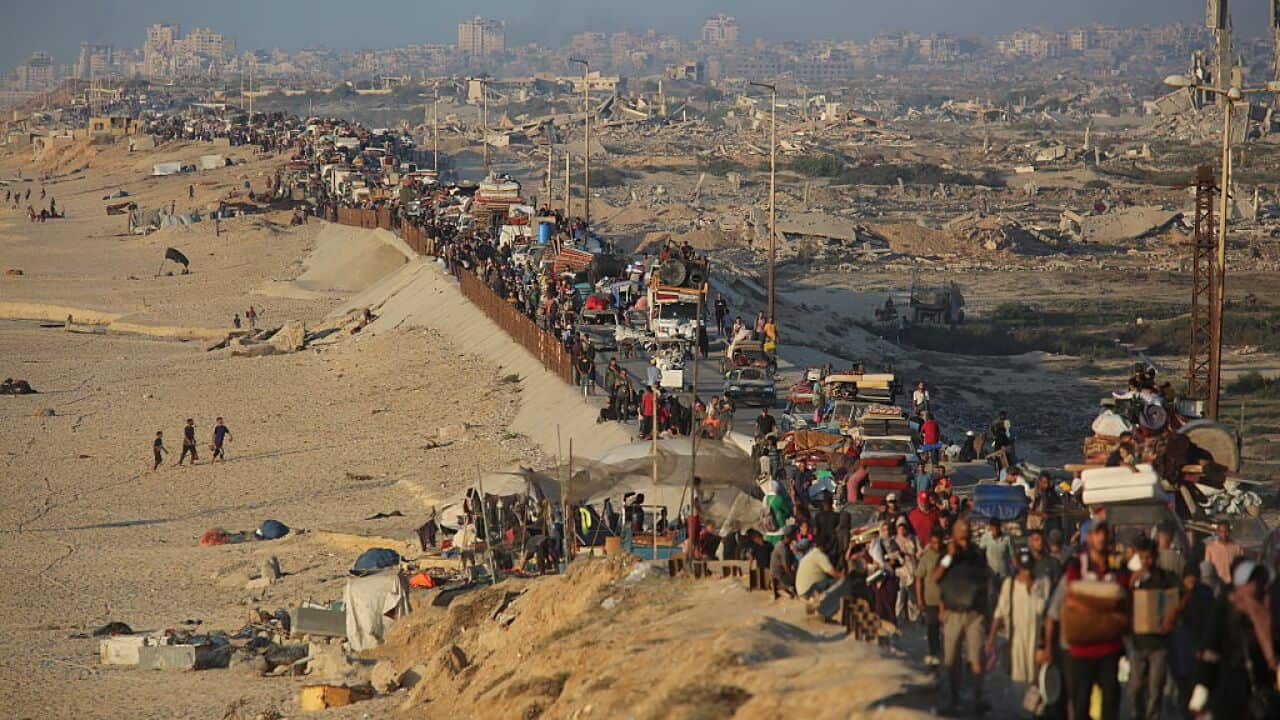 A stream of people carrying belongings walks along a road beside a beach away from a destroyed city.