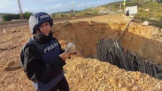 A woman in dark, armoured clothing and a helmet stands near a bomb crater. The word PRESS is emblazoned on her chest and helmet.