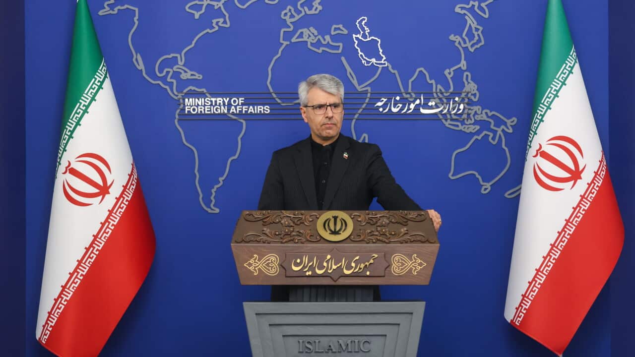 A man with grey hair in black suit stands behind a lectern, in between two Iranian flags.