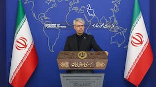 A man with grey hair in black suit stands behind a lectern, in between two Iranian flags.