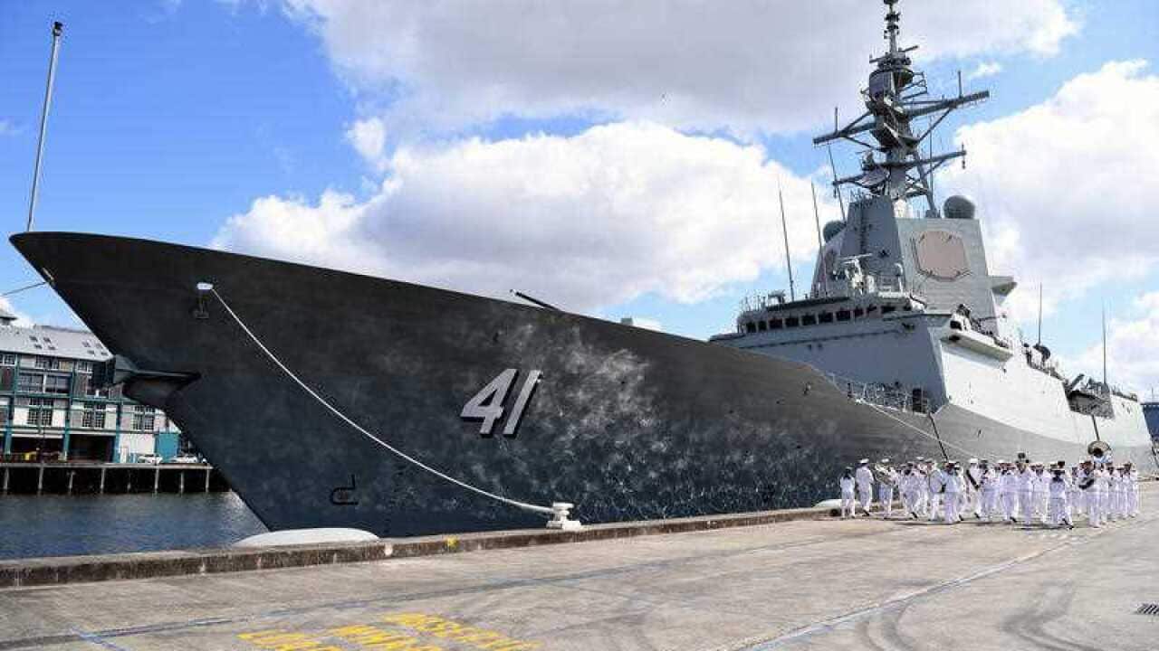 Navy personnel parade alongside a large navy ship tied to a dock. 
