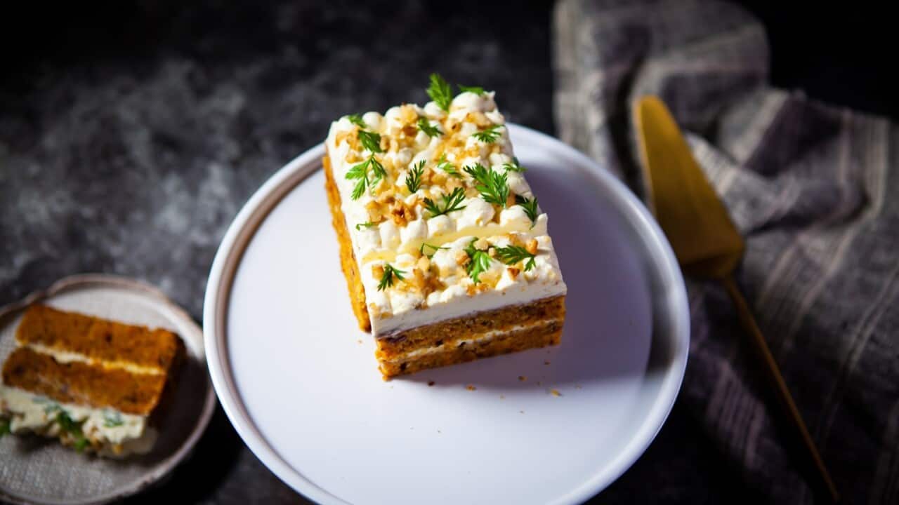 Carrot cake with a thick layer of cream cheese frosting sits on a flat white plate. A slice sits on a plate to one side.
