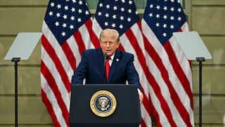 Donald Trump speaking at a platform in front of US flags.