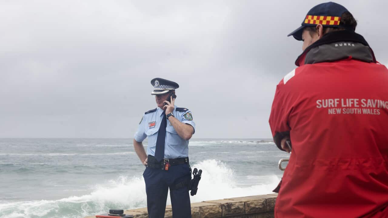 A police officer in a light blue uniform speaks on a mobile phone near a stone wall overlooking a rough ocean, while a person in a red Surf Life Saving New South Wales jacket watches from the foreground.