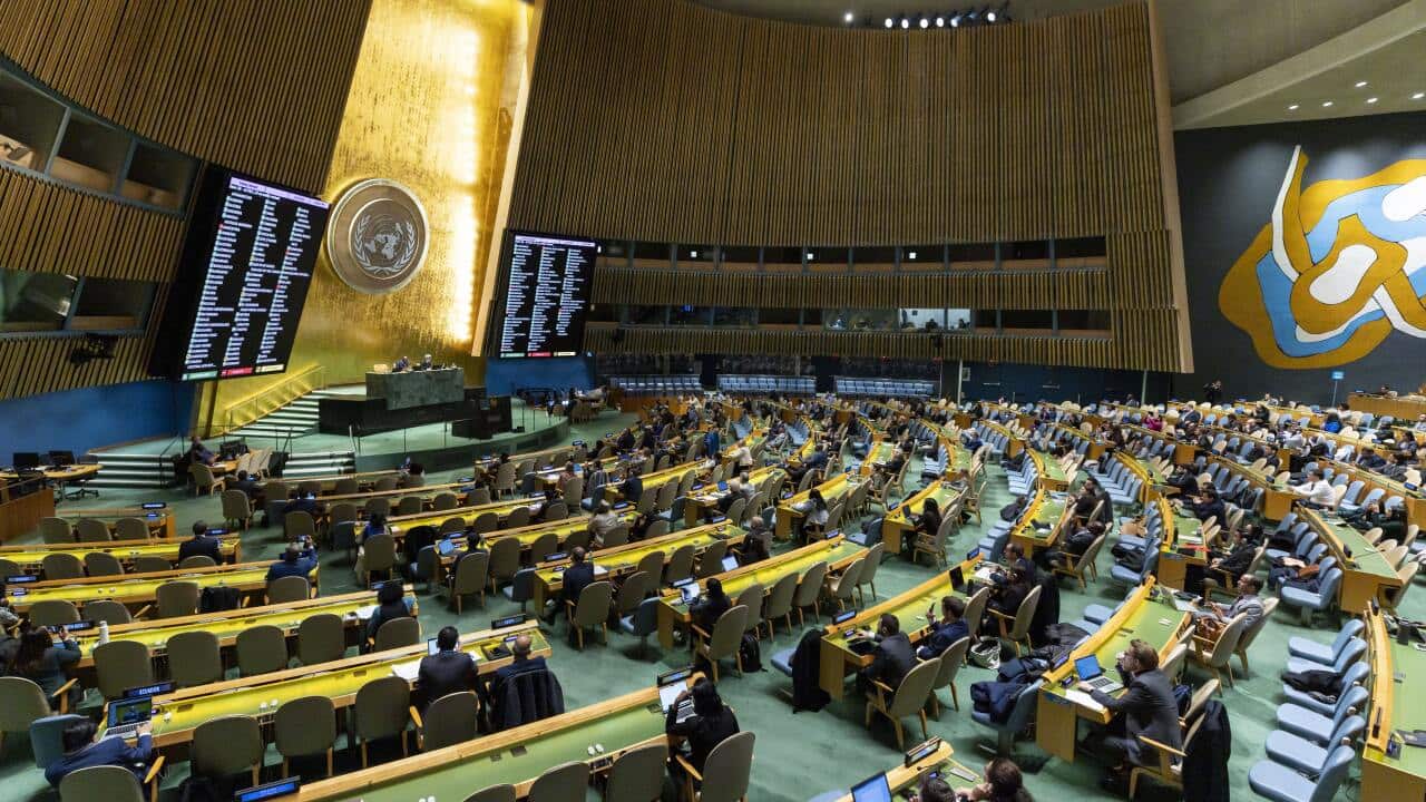 A large group of people sitting at desks in a large hall. There are large screens mounted to the walls.