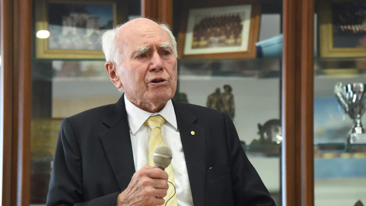 Former prime minister John Howard, stands at a wooden lectern, holding a microphone in his hand as he speaks.