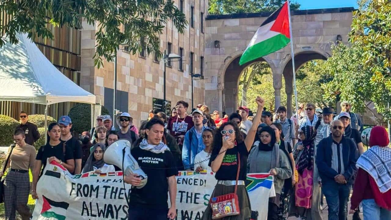 A protest march is taking place on a sunny day, with a crowd of people walking down a path. A woman in the foreground is raising her fist and holding a megaphone, and a large Palestinian flag is visible in the background.