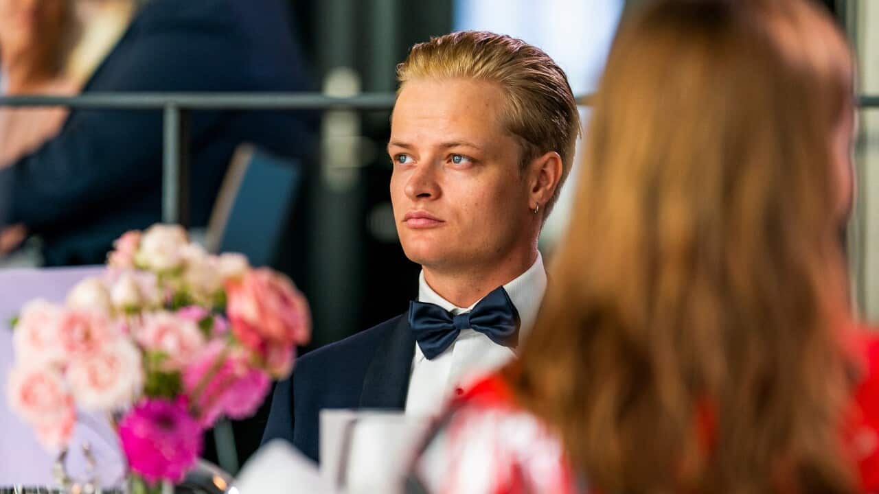 A young man in a black suit and bow tie is looking sideways. There's a woman in a red dress and blonde hair in the foreground with her back turned.