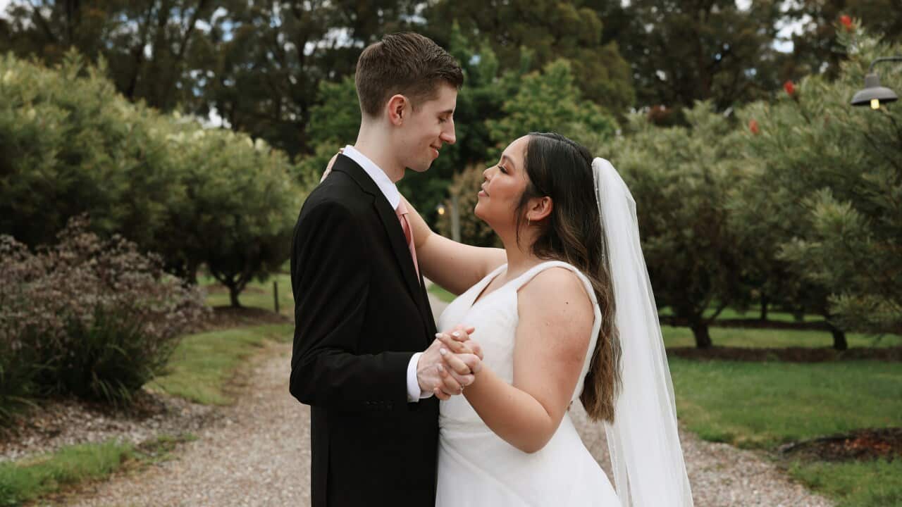 A bride wearing a white dress and a groom in a dark suit hold hands in dance formation, looking at each other lovingly, standing in beautiful garden.