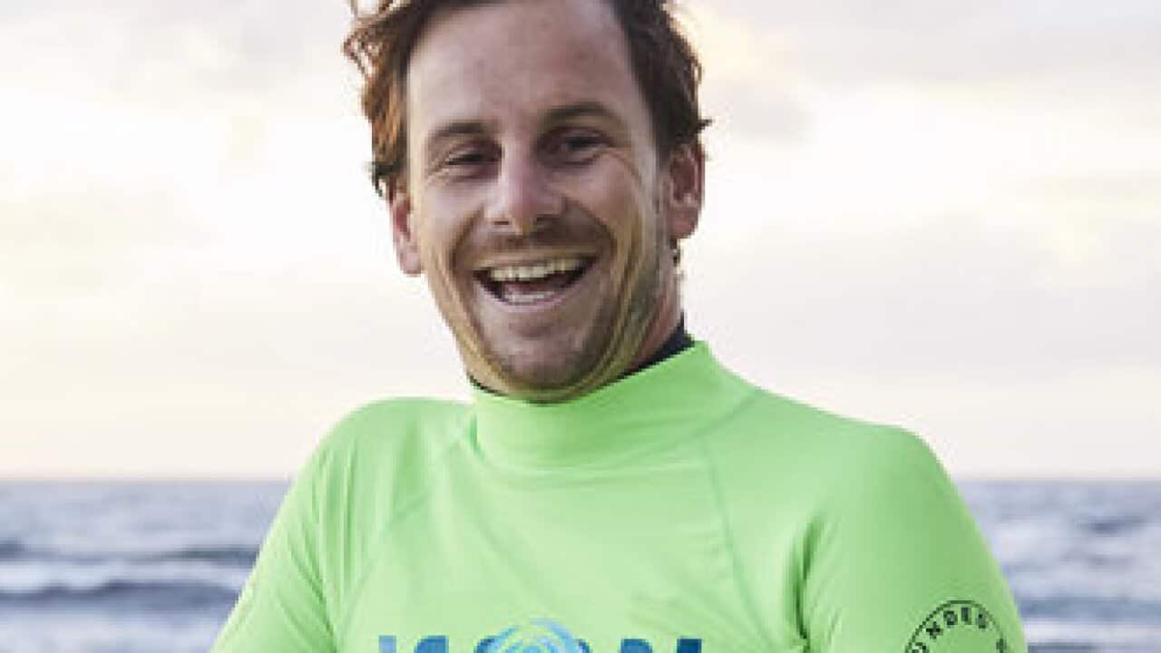 A man who is smiling leaning against a barrier. The ocean is behind him.