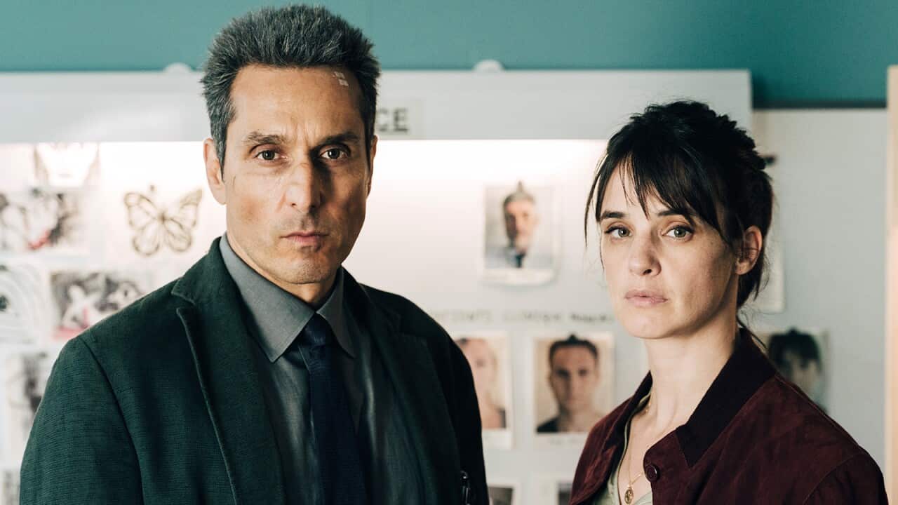 A man and a woman stand in front of a board where several headshots and other pieces of paper have been pinned. They stare intently towards the camera.