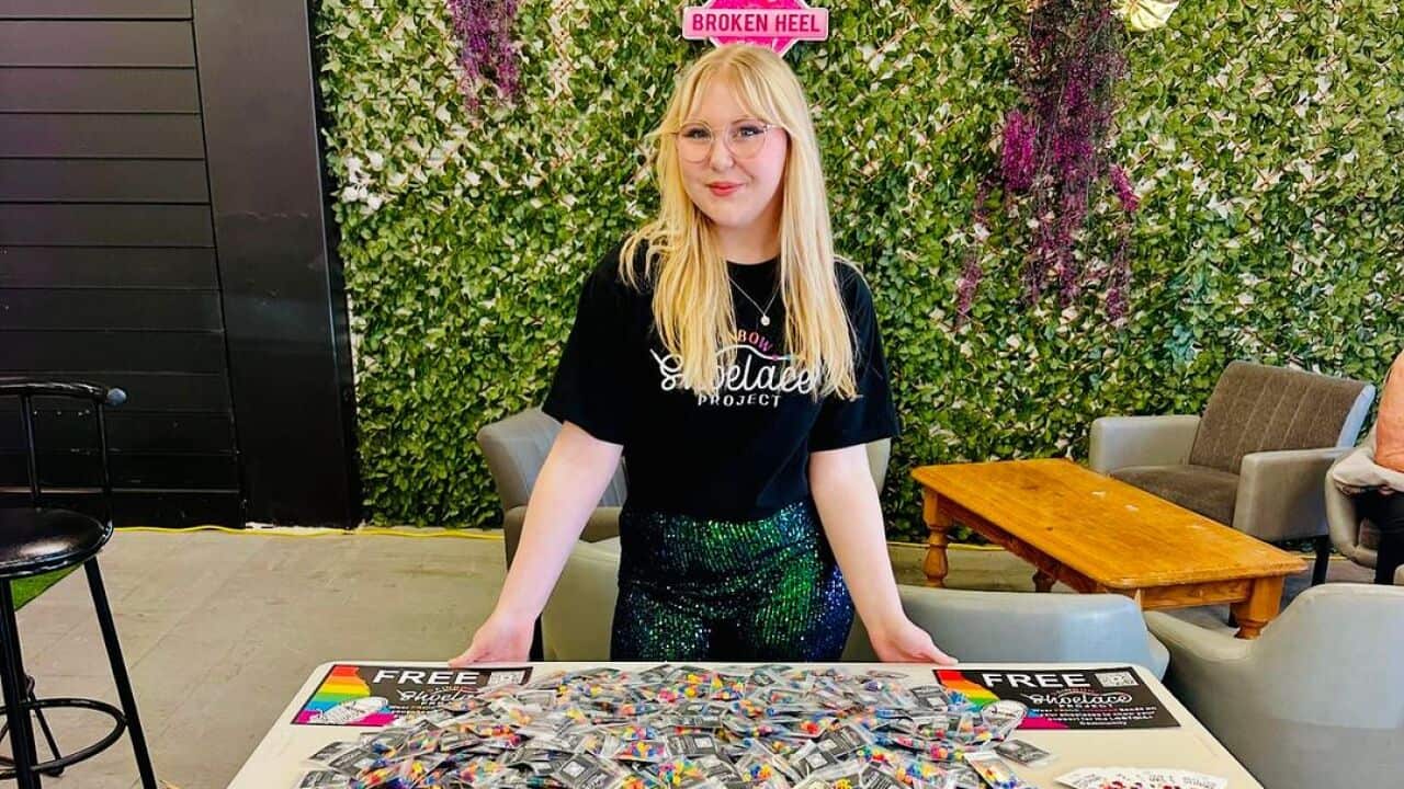 A young blonde girl smiles as she leans across a table with a sign that reads "rainbow shoelace project" littered with little cards