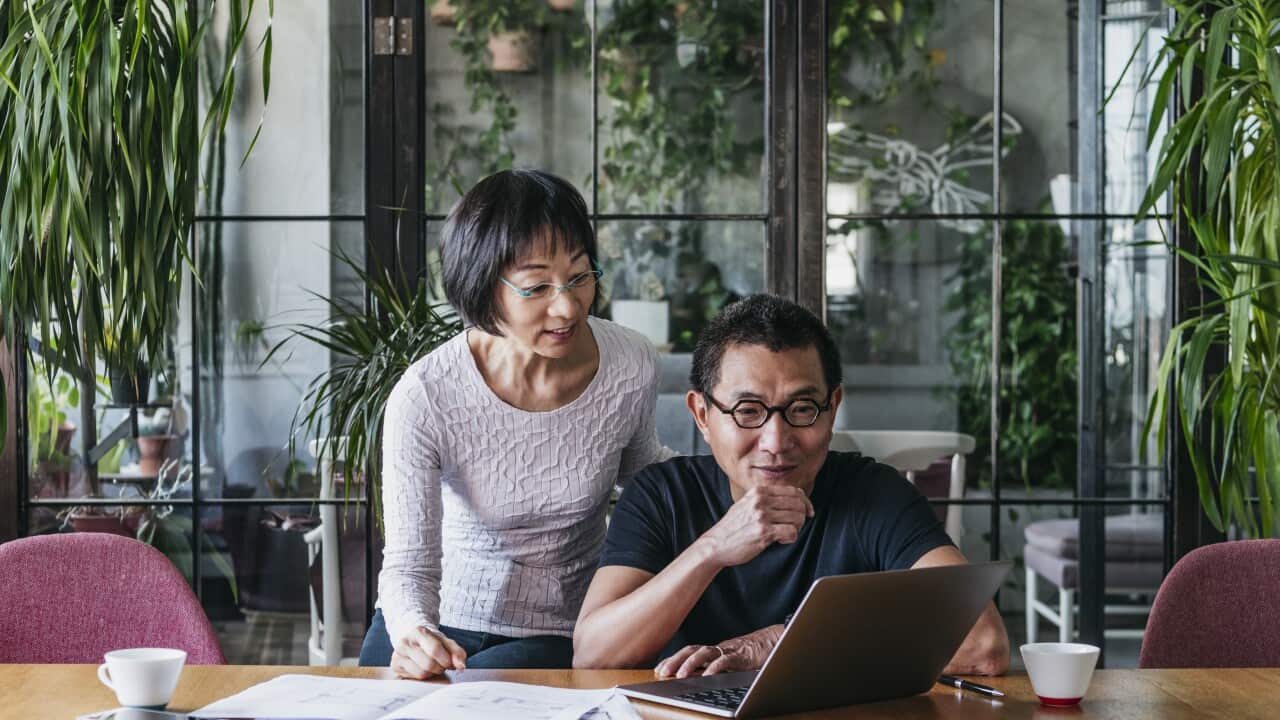 A middle aged man using laptop with a woman looking over shoulder and documents spread on the table.
